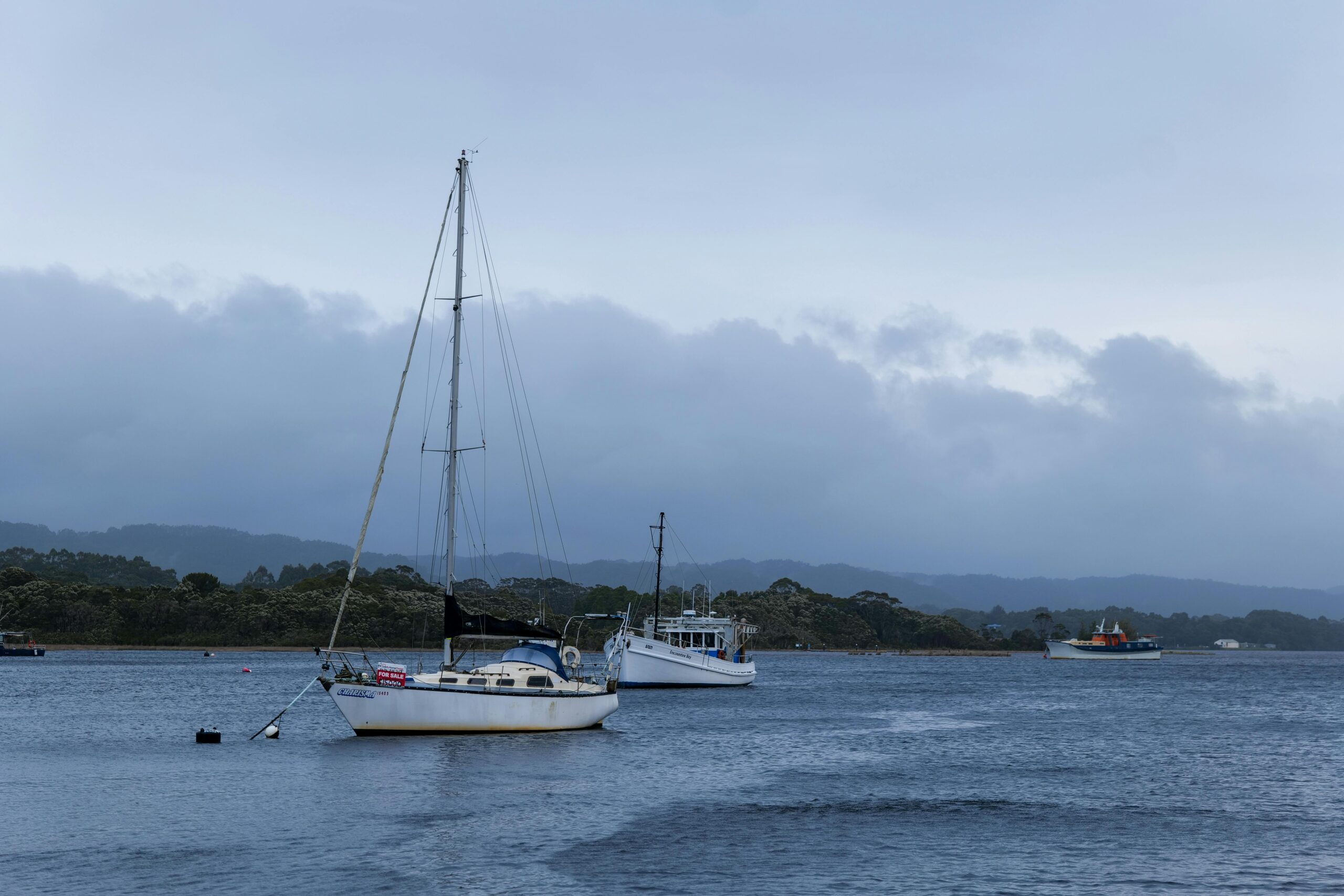 A couple of used boats anchored on calm water under a cloudy sky, with mountains and shoreline in the background.